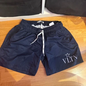 Mörkblå badshorts från Valentino - Snygga mörkblå badshorts från Valentino med VLTN-tryck och stjärna på benet. De har elastisk midja med vit snörning och meshfoder på insidan. Perfekta för stranden eller poolen, med en clean och modern look.