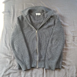 Grå ribbad kofta från East West - Säljer en grå ribbstickad kofta från East West i storlek M. Koftan har hel dragkedja framtill, hög krage och långa ärmar. Materialet är mjukt och stickat, perfekt för lager på lager under höst och vinter. Enkel och clean look som funkar till jeans eller dressade byxor.