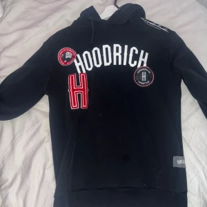 Svart hoodie från Hoodrich med tryck - Svart hoodie från Hoodrich med stora vita och röda tryck på bröstet, samt broderade patches. Tröjan har huva med snörning och en klassisk känguruficka framtill. Långärmad och med ribbade muddar. Perfekt för streetwear-stilen.