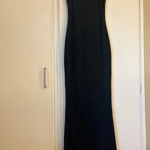 Svart långklänning med smala axelband - En stilren svart långklänning med smala axelband och rak siluett. Klänningen har en enkel design utan mönster eller detaljer, vilket gör den lätt att styla. Perfekt för dig som gillar minimalistiskt och tidlöst mode.