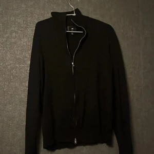 Svart zip-kofta slim fit - Svart kofta från H&M i slim fit-modell med hel dragkedja framtill. Tröjan har lång ärm och en enkel, stilren design som passar perfekt till jeans eller chinos. Materialet är mjukt och bekvämt, och koftan har en hög krage för extra stilpoäng.