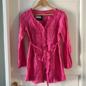 Rosa broderad Odd Molly tunika - Supersöt rosa blus/tunika från Odd Molly med broderade detaljer och spetskant vid halsen. Klänningen har långärmade ärmar, knappar framtill och ett knytband i midjan. På ryggen finns ett broderat citat. Perfekt för dig som gillar färg och unika detaljer!