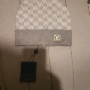 Grå rutig mössa från Louis Vuitton - Snygg grå mössa från Louis Vuitton med klassiskt rutigt mönster och uppvikt kant. Mössan har en metallbricka med LV-logga framtill och är tillverkad i mjukt material som känns lyxigt. Perfekt för dig som vill ha en stilren accessoar med designer-vibe.