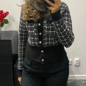 Svartvit rutig kofta med guldfärgade knappar - Snygg svartvit rutig kofta med guldfärgade knappar framtill och två dekorativa bröstfickor. Koftan har ribbade muddar och midja, samt långärmad design. Perfekt för dig som gillar en chic och trendig look med lite edge.