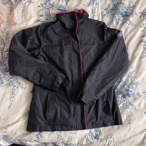 Svart vindjacka med rosa detaljer - Svart vindjacka från Dechatlon med snygga rosa detaljer och foder. Jackan har hög krage, dragkedja framtill och två fickor med dragkedja. Insidan är fodrad i stark rosa färg som ger en cool kontrast. Perfekt för blåsiga dagar och outdoor.