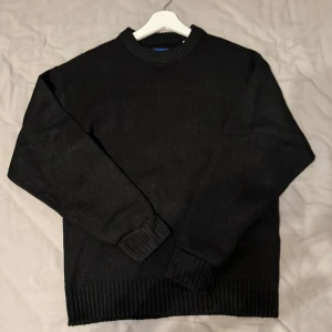 Svart stickad tröja med rund hals - En enkel svart stickad tröja med rund hals och ribbade muddar vid ärmslut och nederkant. Perfekt för dig som gillar stilrena och klassiska plagg. Långärmad och mjuk, passar till jeans eller cargo för en chill look.