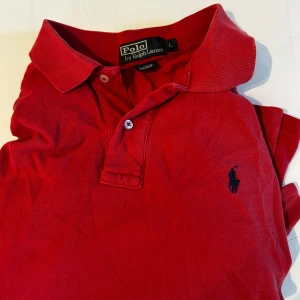 Röd Polo t-shirt från Ralph Lauren - Säljer en klassisk röd Polo t-shirt från Ralph Lauren i storlek L. T-shirten har korta ärmar, krage och två knappar framtill. Materialet är 100% bomull och den har den ikoniska broderade Polo-loggan på bröstet. Perfekt för dig som gillar stilren och tidlös design.