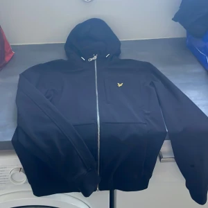 Svart vindjacka från Lyle & Scott - Svart vindjacka från Lyle & Scott med huva och hel dragkedja framtill. Jackan har ett gult broderat fågelmärke på bröstet och praktiska fickor. Materialet är syntet och jackan har en clean, sportig look som passar perfekt till streetwear. Storleken är xs men passar även s