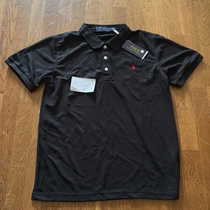 Svart Polo Ralph Lauren t-shirt S - Svart t-shirt från Polo Ralph Lauren i klassisk pikéstil med krage och tre vita knappar framtill. Röd broderad logga på bröstet. T-shirten är kortärmad och gjord i mjuk bomull som känns skön mot huden. Perfekt för en clean och stilren look.
