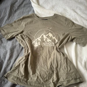 Beige t-shirt med bergtryck - Snygg militär grön t-shirt med coolt vitt tryck av berg, sol och texten 'Adventure is calling'. Rund hals och korta ärmar, perfekt för dig som gillar natur och äventyr. Skön passform och mjukt material som känns chill hela dagen.