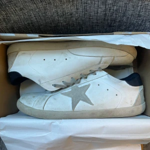Golden Goose Superstar sneakers, vit - Golden Goose Superstar sneakers i vitt skinn med grå mockadetaljer och en ikonisk stjärna på sidan. Skorna har grå snörning, svart häl och rund tå. Snygg, clean look med platt sula och cool branding på innersulan. Perfekta för dig som gillar italiensk streetstyle.