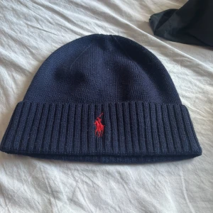 Marinblå ullmössa Polo Ralph Lauren - Snygg marinblå mössa från Polo Ralph Lauren i 100% ull. Klassisk ribbstickad kant och ikonisk röd broderad logga framtill. Perfekt för dig som vill ha en stilren och varm mössa med premiumkänsla och diskret design. Använd max 5 gånger nypris runt 1000kr