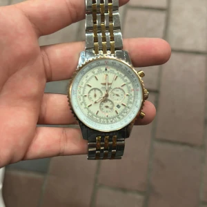 Breitling Navitimer kronograf herrklocka - Snygg Breitling Navitimer herrklocka med kronograf och datum. Armbandet är i silver och guldtonad metall med klassisk länkdesign. Urtavlan är vit med flera subdials och detaljerade markeringar. Guldiga knappar och bezel ger en lyxig känsla. Perfekt för dig som gillar statement-accessoarer.