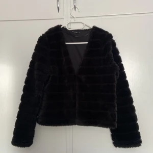 Svart pälsjacka från Gina Tricot - Mjuk och fluffig svart pälsjacka från Gina Tricot i storlek S. Jackan har en rak, kortare passform med markerad v-ringning och långa ärmar. Snygga horisontella ränder i pälsen ger en cool textur. Perfekt för dig som vill ha en trendig look med extra mysfaktor.