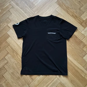 Svart Chrome Hearts t-shirt med tryck - Svart t-shirt från Chrome Hearts i bomull med korta ärmar. På bröstet finns en ficka med Chrome Hearts-logga och på ryggen ett stort, detaljerat vitt kors-tryck. Extra logga på ärmen och nedtill på ryggen. Snygg och clean design med street vibe. (3j äkt4)