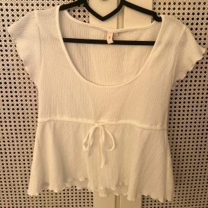 Vit ribbad blus med knyt i midjan - Supersöt vit topp från Nelly med ribbad struktur och volangkant. Storlek S. Använd ett fåtal gånger Den har korta ärmar, rundad halsringning och ett knytband i midjan som ger en extra gullig touch. Perfekt för dig som gillar en feminin och chill stil.