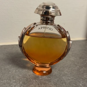 Olympéa parfym från Paco Rabanne - Säljer en Olympéa parfym från Paco Rabanne med en rund, lyxig flaska i transparent glas och roséguldfärgade detaljer. Parfymen har en varm bärnstensfärgad vätska och flaskan pryds av eleganta bladmotiv runt kanterna. Perfekt för dig som gillar exklusiva dofter.