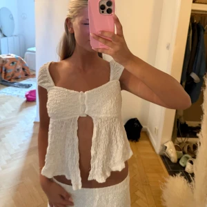 Vit offshoulder blus med volang - Supersöt vit blus med offshoulder och breda axelband. Toppen har en smockad struktur, volangig nederkant och en öppen front med knyt framtill. Perfekt för sommarens alla tillfällen och ger en romantisk vibe.