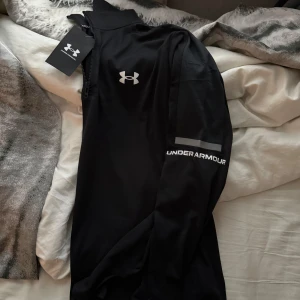 Under Armour  - Svart långärmad träningströja från Under Armour med half zip och logga på bröstet samt ärmen. Snygg sportig look med reflekterande detalj på ärmen. Perfekt för gymmet eller löprundan, tillverkad i mjukt syntetmaterial som andas.