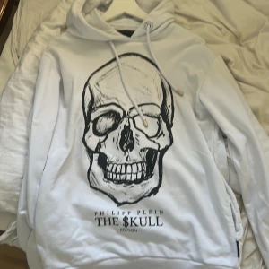 Vit hoodie med döskalle från Philipp Plein - Säljer en vit hoodie från Philipp Plein med stor svart döskalle-tryck på framsidan och texten THE $KULL Edition. Hoodien har snörning i huvan, långa ärmar och en mjuk känsla. Perfekt för dig som gillar edgy streetwear och vill sticka ut. Den passar M också