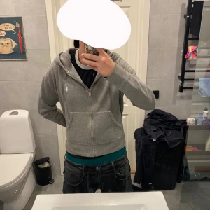Grå Ralph lauren zip hoodie - En grå Ralph lauren zip hoodie. Bra kvalitet knappt använt. Tröjan har broderad logga på bröstet, två fickor framtill och mjukt foder på insidan. Perfekt för chill dagar och enkel att matcha med jeans eller joggers. Storlek L men skulle passar bra om man har storlek M också.
