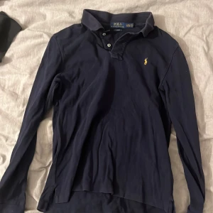 Marinblå långärmad pikétröja Polo Ralph Lauren 18-20 år (passar XS) - Snygg marinblå långärmad pikétröja från Polo Ralph Lauren i slim fit. Klassisk krage och knäppning framtill, med den ikoniska gula broderade loggan på bröstet. Mjuk bomullskvalitet som är skön mot huden och passar perfekt till jeans eller chinos. Passar XS & S