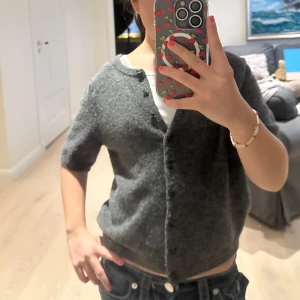 Grå stickad kofta med kort ärm - Mysig grå stickad kofta med korta ärmar och rund hals. Koftan har knappar framtill och en loose passform som ger en chill vibe. Perfekt att slänga över en t-shirt för en enkel och snygg look. Passar dig som gillar basic med en twist.