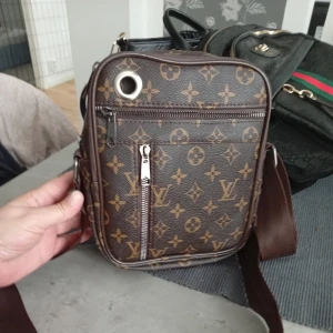 Crossbody väska - Säljer en snygg brun crossbody väska  med klassiskt monogrammönster i beige. Väskan har två dragkedjefack framtill och en bred, justerbar axelrem. Perfekt för att bära med sig det viktigaste och samtidigt hålla stilen.