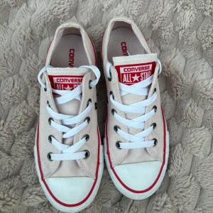 Converse All Star ljusrosa sneakers stl.32 NYA - Säljer ett par klassiska NYA  Converse All Star sneakers i ljusrosa canvas med vit tå och sula, samt röda detaljer runt sulan och på loggan. Skorna har vita snören, metallfärgade öljetter och rund tå. Perfekt för dig som gillar tidlös streetstil. Stl.32 innermått; 19,5 cm