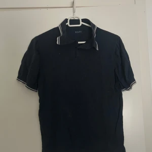 Mörkblå pikétröja från Riley - Snygg mörkblå pikétröja från Riley med vit kant på krage och ärmslut. Klassisk passform och korta ärmar, perfekt för en chill och stilren look. Materialet känns mjukt och bekvämt mot huden. Nypris 1100kr