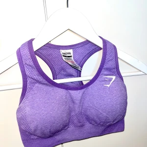 Lila linnetopp från Gymshark XS - Sportig lila linnetopp från Gymshark i storlek XS. Toppen har en snygg racerback-design och är gjord i ett stretchigt syntetmaterial med mesh-detaljer för extra ventilation. Perfekt för träning och gym, med vit logga framtill och bak.