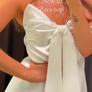 Vit glättad  topp med knytning  - Supersnygg vit glättad  topp från Zara i glansigt satinmaterial. Toppen har en stor rosett framtill som ger en extra wow-effekt och är axelbandslös för en chic look. Perfekt för dig som vill sticka ut och älskar trendiga detaljer. Prislapp kvar och aldidg använd förut. Slutsåld på Zaras hemsida 