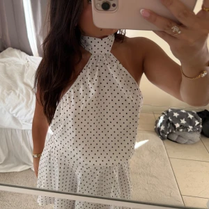 Vit prickig kortklänning med halterneck - Superfin vit kortklänning med svarta prickar och halterneck. Klänningen har en öppen rygg och ett flowigt, volangigt kjolparti som ger en skön och luftig känsla. Perfekt för varma dagar och när du vill sticka ut med en söt och trendig look.