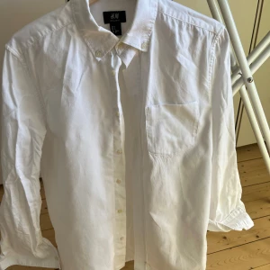 Vit skjorta från H&M Regular Fit - Klassisk vit skjorta från H&M i regular fit med knappar hela vägen och en bröstficka. Skjortan har lång ärm och krage med knappdetaljer. Perfekt för dig som gillar enkel och stilren look.