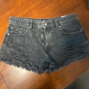 Svarta jeansshorts från Sinsay - Coola svarta jeansshorts från Sinsay i storlek 38. De har en högmidjad passform, slitna detaljer och fransig kant nedtill för en edgy vibe. Klassisk femficksmodell med knapp och dragkedja framtill. Perfekta för sommarens alla äventyr.