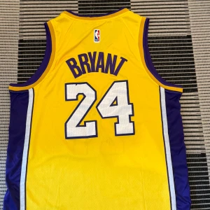 Lakers Kobe Bryant #24 baskettröja - Säljer en ikonisk Lakers baskettröja med Kobe Bryant #24. Tröjan är ärmlös och har en stark gul färg med lila och vita detaljer längs sidorna. Lakers-logga på framsidan, Bryant och nummer 24 på ryggen. Materialet är mesh för bra ventilation, perfekt för sport eller streetwear.