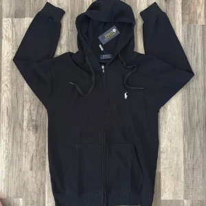 Svart hoodie från Polo Ralph Lauren - Svart hoodie från Polo Ralph Lauren med dragkedja och huva. Klassisk design med vit broderad logga på bröstet, snörning i huvan och två fickor framtill. Tillverkad i mjuk bomull som är skön att ha på sig. Perfekt för chill dagar eller när du vill ha en clean look.