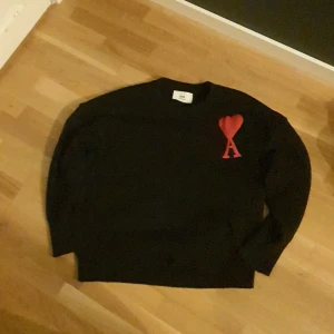 Svart stickad tröja från Ami - Svart stickad tröja från Ami Alexandre Mattiussi med lång ärm och ribbade muddar. På bröstet finns ett rött hjärta med bokstaven A, vilket ger en unik och snygg detalj. Perfekt för dig som gillar stilrena plagg med en twist. 