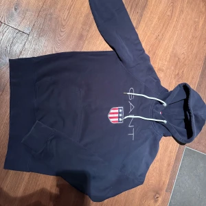 Mörkblå hoodie från GANT med emblem - Snygg mörkblå hoodie i mjuk bomull från GANT med broderat emblem och vita snören i huvan. Tröjan har lång ärm, klassisk känguruficka och GANT-logga på bröstet. 