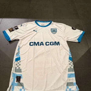 Marseille fotbollströja Puma Dedić 77 - Snygg vit Marseille fotbollströja från Puma med blå detaljer och tryck. Kortärmad med blå krage och ärmslut, samt rutmönster längs sidorna. Framsidan har CMA CGM-logga och klubbmärke, baksidan har Dedić och nummer 77 i blått. Materialet är lätt och andas, perfekt för match eller träning.