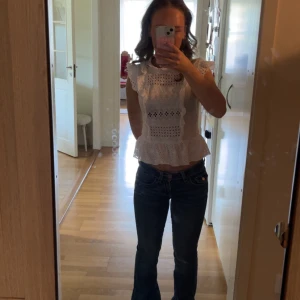 Vit blus med volang och broderi - Säljer en söt vit blus med volangkant och broderade detaljer. Blusen har ärmlös design med rysch vid axlarna och ett genombrutet mönster framtill. Passar perfekt till både jeans och ljusa byxor. Supersnygg till vår och sommar!