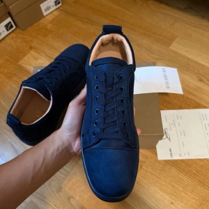 Mörkblå sneakers från Louboutin storlek 42 - Snygga mörkblå sneakers från Louboutin med klassisk röd sula och diskret broderad logga på hälen. Skorna har rund tå, platt sula och är tillverkade i mocka med snörning framtill. Insidan är ljusbeige och ger en lyxig känsla. Perfekt för dig som gillar stilrena detaljer.
