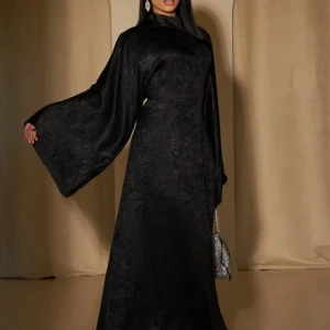 Svart långklänning med vida ärmar - Säljer en svart långklänning med hög hals och riktigt vida, svepande ärmar. Klänningen har en elegant, glansig yta och sitter snyggt med en rak siluett. Perfekt för dig som vill sticka ut med en dramatisk look och maxad stil.