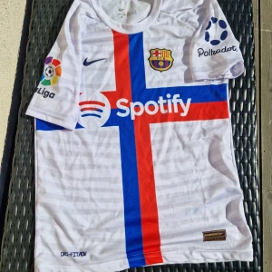 FC Barcelona Ansu Fati Nike fotbollskit - Säljer en FC Barcelona fotbollströja med Ansu Fati och nummer 10 på ryggen. Tröjan är vit med ett stort kors i blått och rött framtill, samt sponsorloggor från Spotify, La Liga och Polkadot. Kortärmad och tillverkad i lätt, ventilerande Dri-FIT-material. Matchande vita shorts med klubbmärke och Nike-logga ingår.