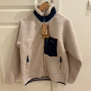 Patagonia Pile retro jacket strl M - Säljer en sjukt mysig vit teddyjacka från Patagonia i storlek M. Jackan har blå detaljer, hög krage, dragkedja framtill och en mörkblå bröstficka. Perfekt för kalla dagar och riktigt skön att ha på sig. Fleece-materialet gör den extra varm och fluffig. Säljer den åt min kille då den var för liten och ej går att lämna tillbaka😊