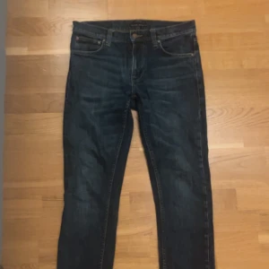 Mörkblå jeans från Nudie - Snygga mörkblå jeans från Nudie med klassisk femficksdesign och gul broderad våg på bakfickorna. Jeansen har en rak passform och är tillverkade i slitstarkt denim. Perfekta för dig som gillar stilrena och tidlösa jeans. Storleken är 28/32.