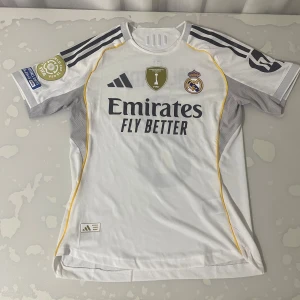 Real Madrid Valverde matchtröja 2024 - Säljer en officiell Real Madrid fotbollströja från Adidas, säsong 2024, med Valverde och nummer 8 på ryggen. Tröjan är vit med grå detaljer, gula linjer och svarta tryck. Slim fit, kortärmad och tillverkad i ventilerande polyester. FIFA Intercontinental Champions-märke på bröstet.