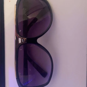 Louis Vuitton svarta solglasögon - Säljer ett par svarta Louis Vuitton solglasögon med stora, rundade bågar och lila tonade glas. Sidorna har guldfärgade detaljer med LV-logga och mönster. Pris kan diskuteras så kom gärna med bud!