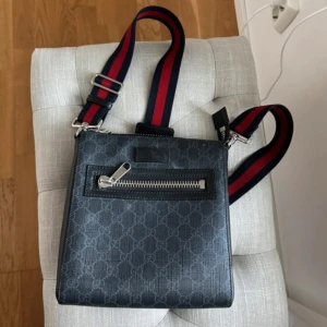 Svart axelväska från Gucci med mönster - Snygg svart axelväska från Gucci med klassiskt GG-mönster och bred justerbar rem i rött och marinblått. Väskan har en praktisk dragkedja framtill och är tillverkad i skinn med silverfärgade detaljer. Perfekt för dig som vill ha en stilren och lyxig look.