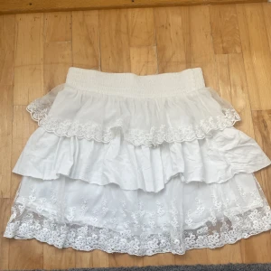 Vit volangkjol med spets från KappAhl - Supersöt vit kjol från Vintage Stories by KappAhl med flera lager volanger och detaljerad spets längst ner. Kjolen har bred resår i midjan och romantiska broderier på tyll. Perfekt för dig som gillar en feminin och drömmig stil.